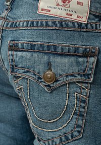 Gros plan de la poche arrière d’un jean en denim bleu avec rabat et bouton, surpiqûres orange et une étiquette blanche avec un texte rouge et noir près de la taille.