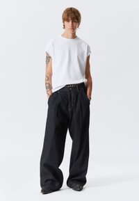 Joven con cabello castaño claro, vistiendo una camiseta sin mangas blanca y unos pantalones vaqueros oscuros de gran tamaño, de pie con las manos en los bolsillos sobre un fondo blanco.
