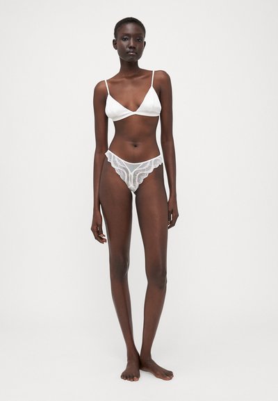 Bralette bianco in pizzo con spalline sottili, abbinato a culotte in pizzo trasparente con un design a bordi ondulati. La modella è in piedi a piedi nudi.