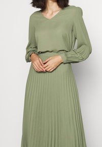 Abito verde oliva con gonna a pieghe, scollatura a V e maniche lunghe arricciate. Realizzato in tessuto leggero, presenta una vestibilità comoda.