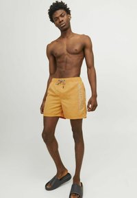 Bañador naranja con cintura elástica y cordón ajustable, con un logo en el lateral. Combinado con chanclas negras.