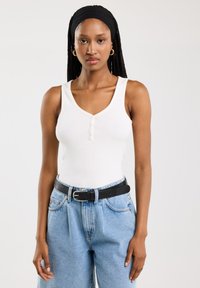 Valge ribakujuline tank top V-kaelusega ja kolme nööbiga, kombineeritud kõrge vöökohtadega helesiniste denim pükstega ja musta vööga.