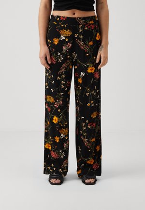 Femme portant un pantalon noir à jambes larges avec un motif floral multicolore et des sandales noires à bout ouvert, debout devant un fond uni.