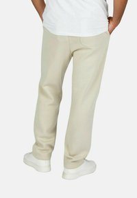 Hellbeige Baumwoll-Sweatpants mit entspannter Passform, ausgestattet mit Gesäßtaschen und Bündchen. Kombiniert mit einem weißen-Shirt und weißen Sneakers.