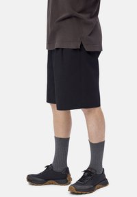 Person wearing black athletic shoes with brown soles, dark gray ribbed socks, black shorts, and a dark brown shirt.