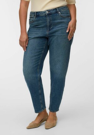 Persona vestida con vaqueros rectos de tiro alto azules y zapatos planos beige, de pie con las manos relajadas a los lados.