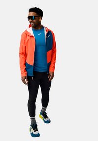 Veste coupe-vent orange vif et bleu marine avec une capuche, une chemise bleue en dessous, des leggings noirs et des chaussures de course colorées avec des semelles texturées.