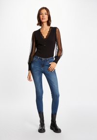 Maglietta nera a maniche lunghe con maniche trasparenti a pois e bordo di pizzo, abbinata a jeans skinny blu e stivaletti neri.