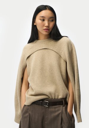 Junge Frau mit langen dunklen Haaren, die einen beigen gestrickten Cape-Pullover, braune Nadelstreifenhosen und einen schwarzen Gürtel trägt, Hände in den Taschen.