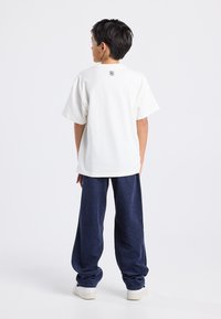 Valkoinen lyhythihainen t-paita, jossa on pyöreä pääntie ja logo takana, yhdistettynä sinisiin korkkiruuvihousuihin ja valkoisiin tennareihin.