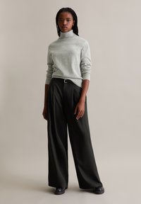 Grauer Rollkragenpullover kombiniert mit weiten schwarzen Hosen und schwarzem Schuhwerk. Der Stoff scheint weich zu sein mit gerippten Akzenten an den Bündchen und dem Saum.