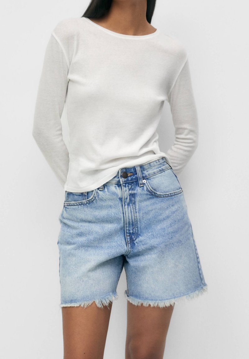 Femme portant un haut blanc ajusté à manches longues et un short en jean bleu clair taille haute avec des ourlets effilochés sur un fond uni.