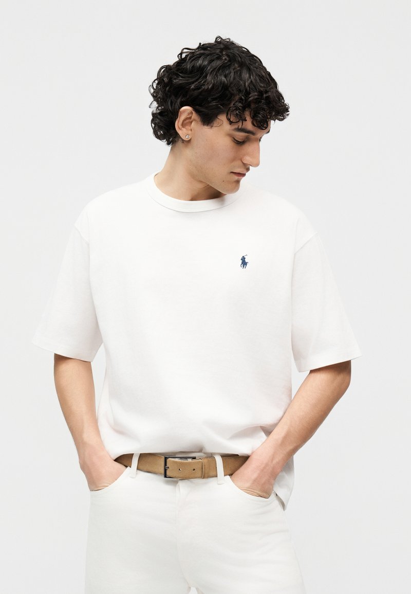 Jonge man met krullend haar, gekleed in een wit shirt met korte mouwen met een blauw logo, witte broek en een beige riem, handen in de zakken, kijkend naar beneden.