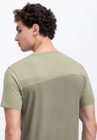 T-shirt verde oliva con texture a gradiente, maniche corte e un piccolo logo a forma di forbici impresso sul colletto posteriore.