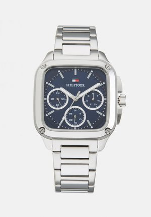 Montre en acier inoxydable argenté avec un cadran carré bleu marine. Les caractéristiques comprennent trois sous-cadrans, des index horaires argentés et un détail de logo.