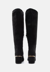Bottes hautes en daim noir avec talons carrés et bordure dorée, vues de dos sur un fond blanc.
