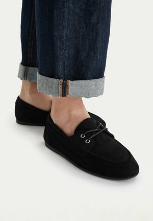 Zapatos mocasín de ante negro con detalle de cordones, usados con jeans de mezclilla azul oscuro remangados sobre pies descalzos.