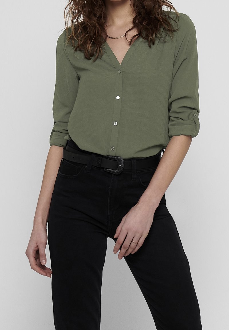 Femme portant une chemise olive à manches remontées, un pantalon noir avec ceinture noire, et un collier simple, debout devant un fond uni.