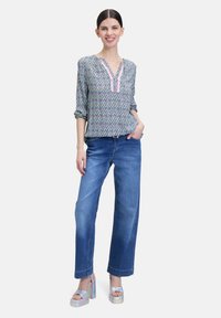 Bunte gemusterte Bluse mit V-Ausschnitt und hochgekrempelten Ärmeln, kombiniert mit lockeren blauen Jeans und schillernden Plateau-Sandalen.