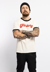 Camiseta cremosa con ribete negro y la palabra "Forsberg" en letras rojas en negrita en el pecho. El modelo tiene tatuajes y lleva un gorro negro.