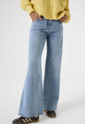 Hellblaue Schlagjeans mit hoher Taille, subtilen Used-Details und fransigem Saum, kombiniert mit dunklen Sneakers und einem gelben Strickpullover.