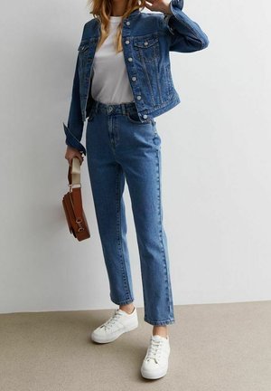 Femme portant une veste en jean bleue et un jean avec une chemise blanche et des baskets, tenant un sac à main marron contre un mur blanc.