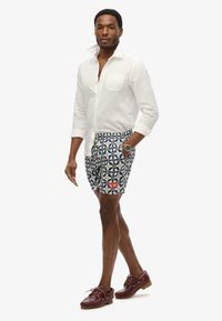 Mężczyzna w białej koszuli zapinanej na guziki, niebiesko-białych szortach w kwiatowy wzór, zegarku na nadgarstku i brązowych skórzanych butach żeglarskich.