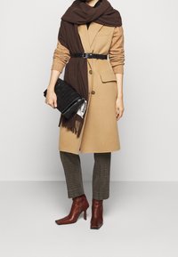 Beige ullkappa med en brun sjal, svart bälte, rutig byxor och burgundy ankelskor. En texturerad svart clutchväska hålls i ena handen.