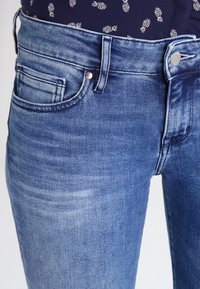Jeans de denim azul claro con una textura suave, que presentan un diseño clásico de cinco bolsillos, un cierre de botón remachado y un ligero desvanecimiento.