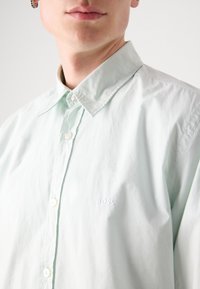Camisa de botones verde claro con cuello clásico. Textura suave con un brillo sutil, que presenta un logo bordado "BOSS" en el pecho.