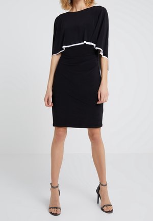 Femme portant une robe noire ajustée longueur genou avec une couche supérieure ample bordée de blanc, associée à des sandales à talons hauts noires avec brides autour de la cheville.