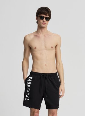 Uomo senza maglietta che indossa pantaloncini da bagno neri con la scritta "TERRANOVA" sulla gamba sinistra e occhiali da sole neri, in piedi davanti a uno sfondo grigio uniforme.