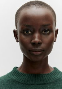Person mit kurzem, strukturiertem Haar, der einen dunkelgrünen, gerippten Pullover trägt. Der Hintergrund ist schlicht und hell, um die Gesichtszüge hervorzuheben.