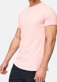 Kurzarm-T-Shirt mit Rundhalsausschnitt aus hellrosa Baumwolle, ausgestattet mit einem kleinen weißen Logo auf der Brust und einem lässigen Schnitt.