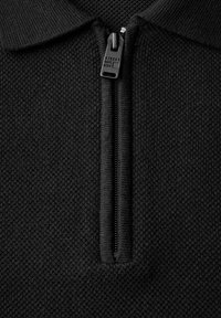 Svart tröja med en strukturerad stickad mönster, med en halv dragkedja framtill och en krage. Dragkedjans dragare i ett kontrasterande material.