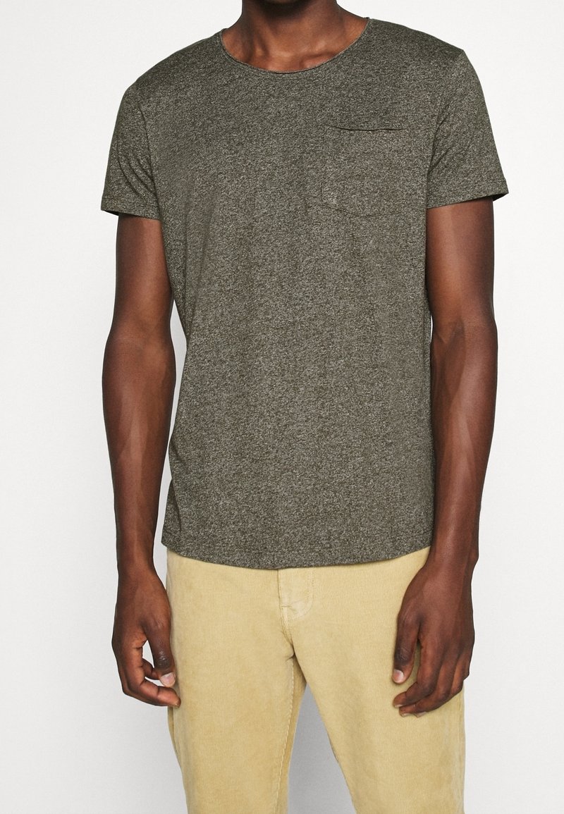 Homme portant un T-shirt texturé vert olive à manches courtes avec une poche poitrine et un pantalon beige clair sur fond uni.