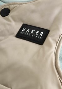 Béžová tkanina s hladkou textúrou, s čiernym obdĺžikovým štítkom, na ktorom je nápis "BAKER BY TED BAKER," doplnená tmavou gombíkom.
