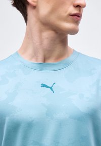 Νέος άνδρας που φοράει ανοιχτό μπλε μπλουζάκι Puma, με εμφανή τον λαιμό και τη γραμμή του σαγονιού, κοιτάζοντας προς τα δεξιά σε ένα απλό φόντο.