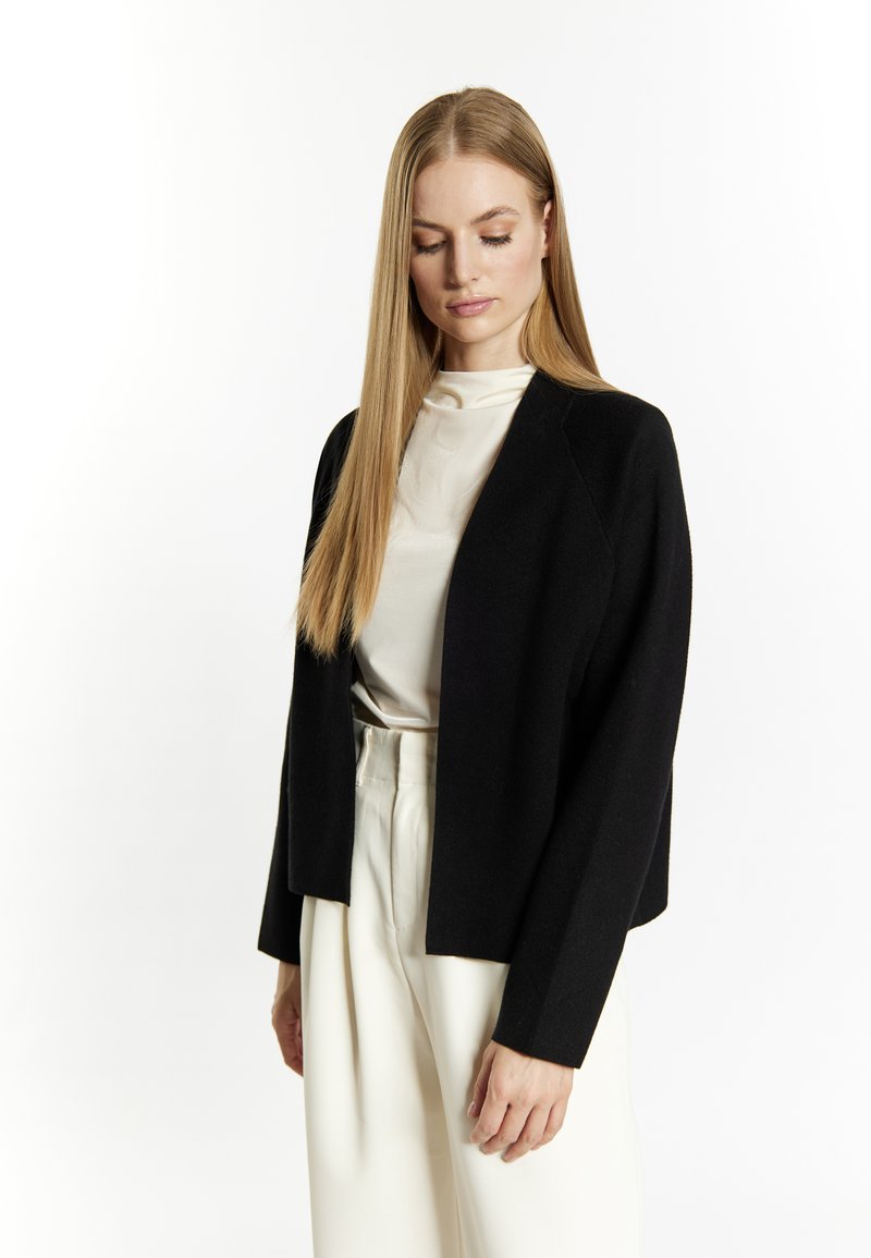 Sort open-front cardigan lavet af blødt materiale, med lange ærmer og cropped design, parret med en lys top og beige bukser.