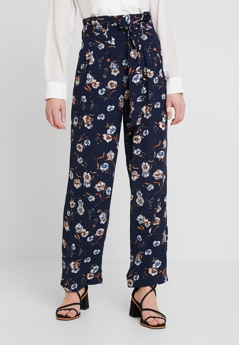 Pantalon large bleu marine à imprimé floral blanc et marron, ceinture nouée, associé à un haut à manches longues blanc et des sandales à talons noires.