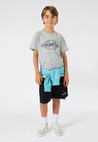 Hall triiksärk "CHAMPION" logoga, mustad lühikesed püksid ja helesinine kampsun vööl seotud. Valged sokid ja tossud täiendavad riietust.