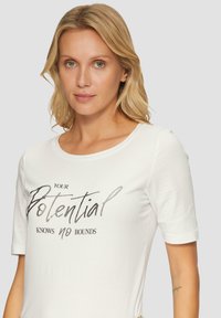 Witte t-shirt met korte mouwen van zachte stof met een bedrukte tekst "JOUW potentieel kent geen grenzen" in zwart en grijs.