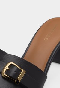 Mule en cuir noir à talon ouvert avec une large lanière, détail de boucle dorée, et semelle intérieure beige, dotée d'un petit talon carré.