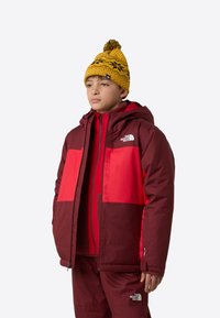 Red and burgundy winter jacket with a hood, featuring color-block design. Paired with a yellow patterned beanie.