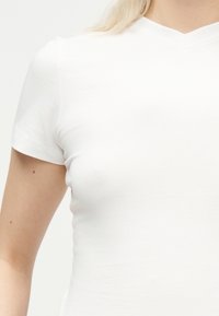 Κλείσιμο εικόνας ενός ατόμου που φοράει απλό λευκό T-shirt με κοντό μανίκι και V-λαιμόκοψη, δείχνοντας τον άνω κορμό και το χέρι του μπροστά σε ανοιχτό φόντο.