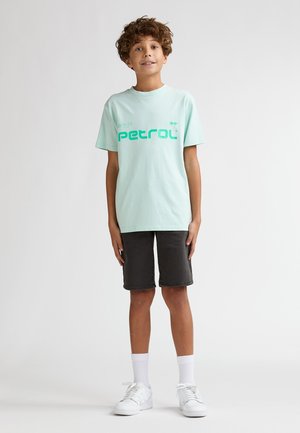 Maglietta a maniche corte verde menta chiaro con una grafica audace "petrol" in turchese. Abbinata a pantaloncini grigio scuro fino al ginocchio e sneaker bianche.