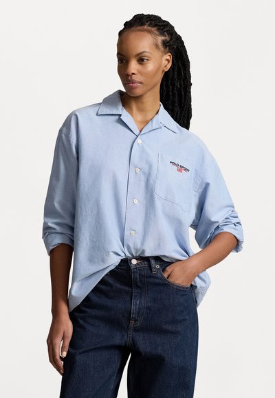 Light blue button-up shirt made of soft material, featuring a pocket with "POLO SPORT" embroidery. Paired with dark denim jeans.