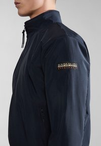 Mørkeblå vindjakke med høj krave, lynlåslomme og et struktureret stof. Har et logo patch på venstre ærme.