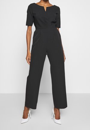 Sort jumpsuit med figursyet overdel, korte ærmer og en keyhole-udskæring. Design med vide ben, tekstureret stof, kombineret med hvide høje hæle.