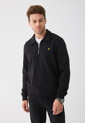 Sweatshirt noir avec une fermeture éclair quart, un col plat et un logo avant en doré. Doté de manches longues et d'une coupe décontractée. Porté avec un pantalon sombre.